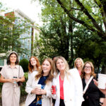 A group of seven women stand outdoors, smiling and holding drinks, with trees and buildings in the background on a bright day.