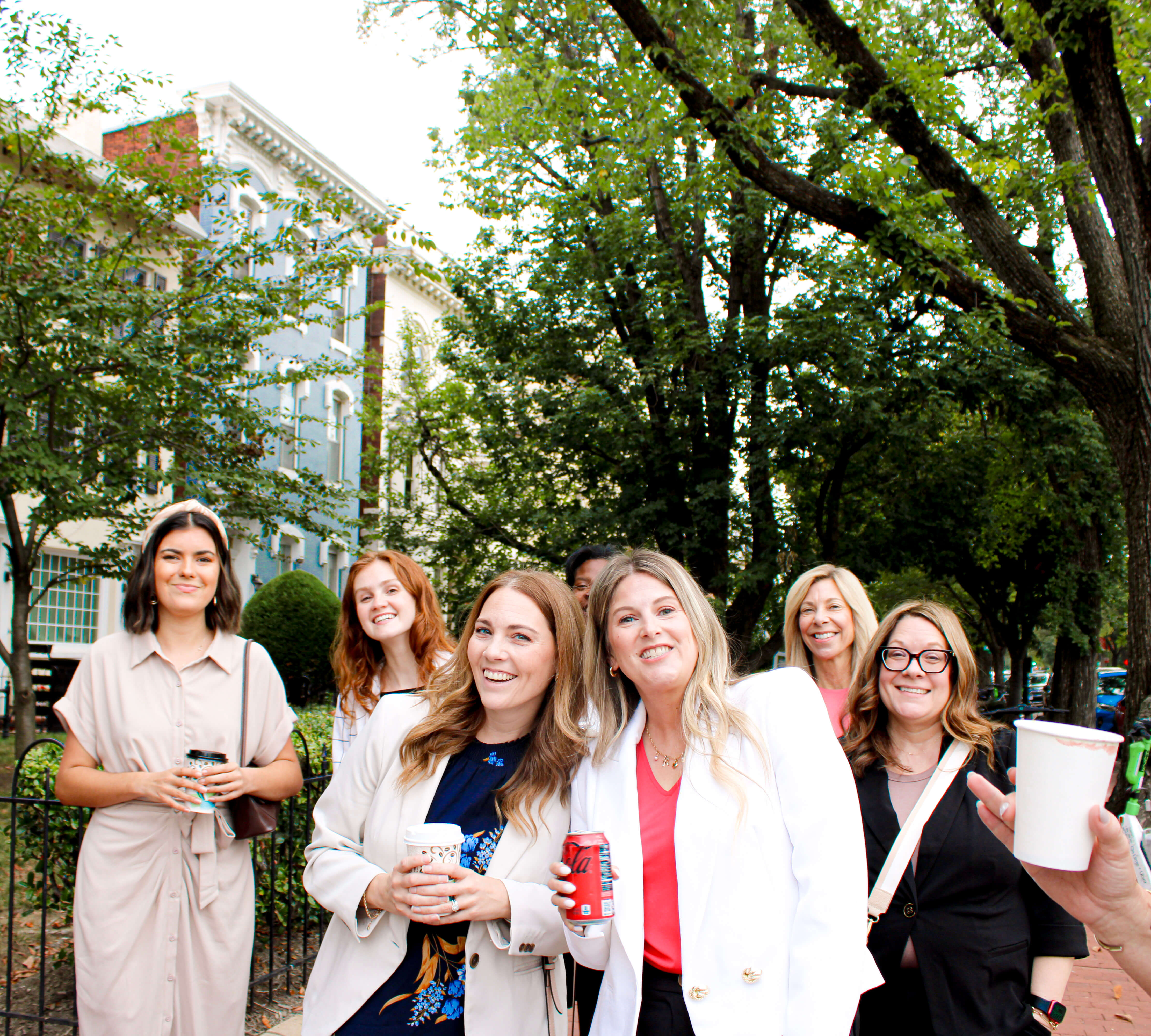 A group of seven women stand outdoors, smiling and holding drinks, with trees and buildings in the background on a bright day.