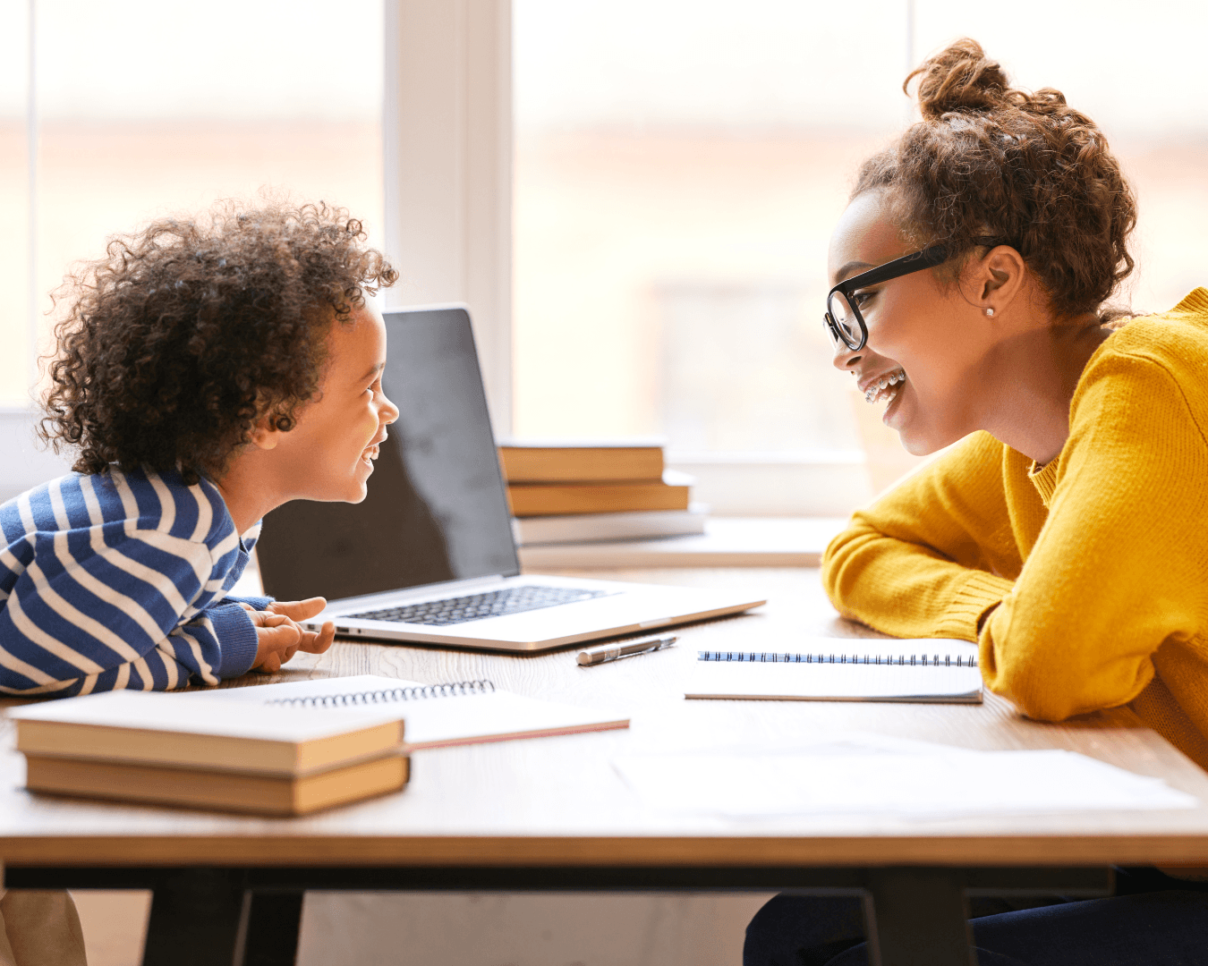A woman and a child sit at a desk smiling at each other. Books, notebooks, and a laptop are on the desk, and sunlight streams through the window behind them.