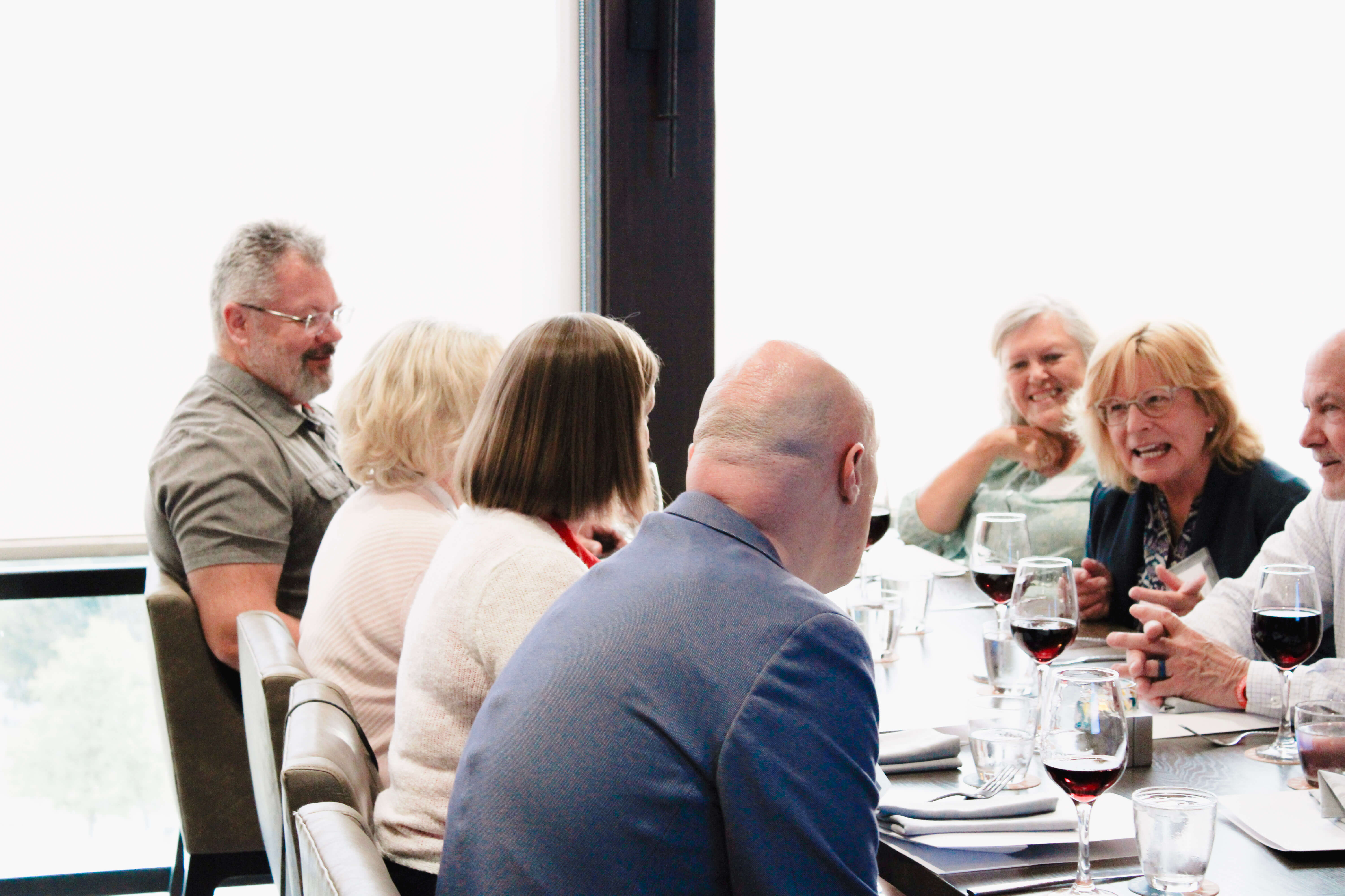 A group of adults sit around a dining table, talking and laughing together. They have wine glasses in front of them, and the atmosphere appears lively and friendly.
