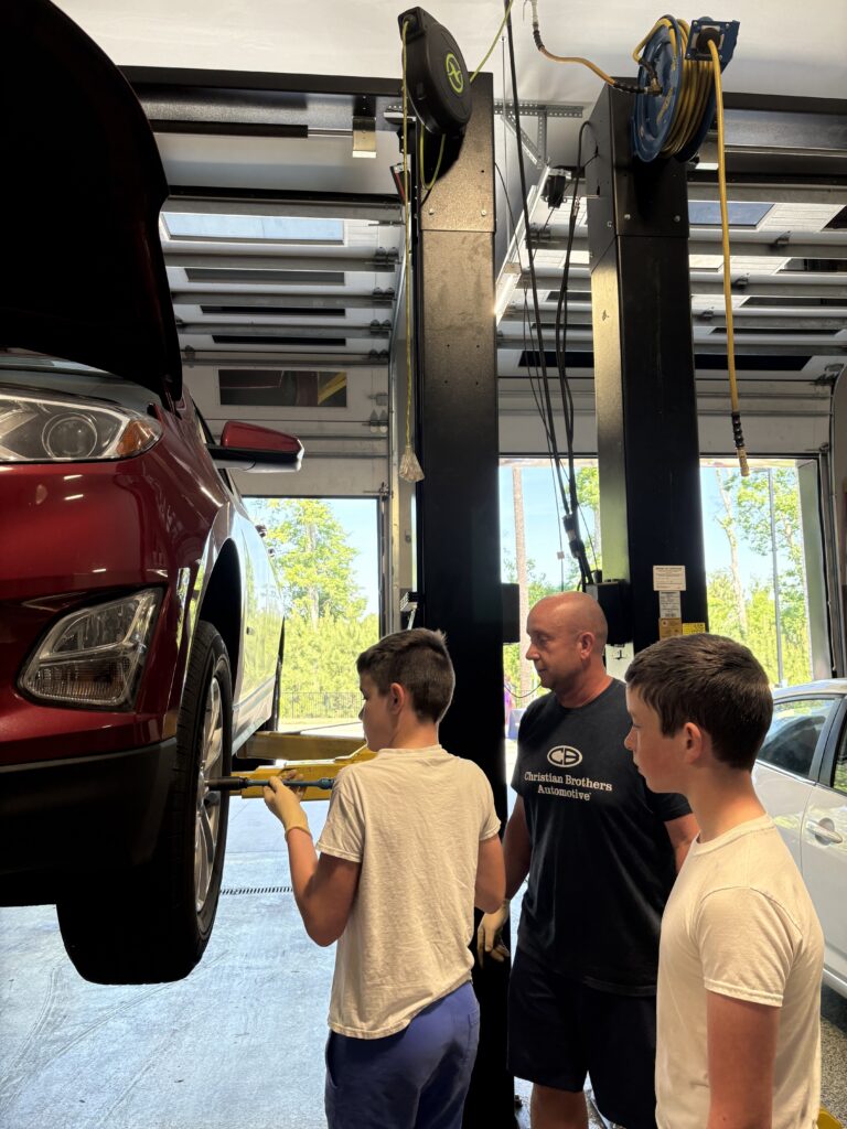 Two boys and an adult stand beside a red SUV raised on a lift in an auto repair shop. The adult supervises as one boy inspects the cars wheel while the other observes. Sunlight shines through large windows behind them.