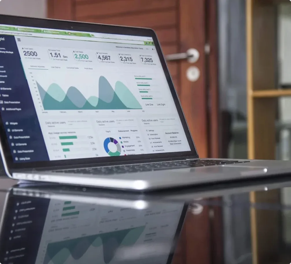 A laptop sits on a glossy table displaying data analytics dashboards with graphs, charts, and statistics. The background features a wooden door and shelves, creating a modern office setting.