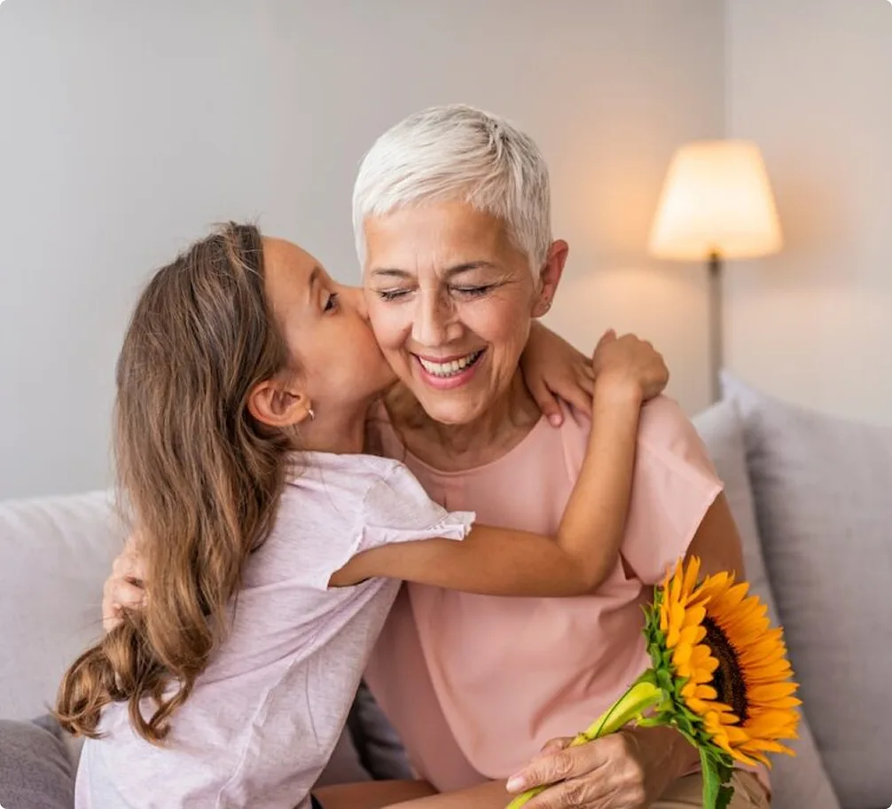 A smiling older woman with short gray hair hugs a young girl who is kissing her cheek. The woman holds a sunflower bouquet, and they are sitting on a couch with a lamp in the background.