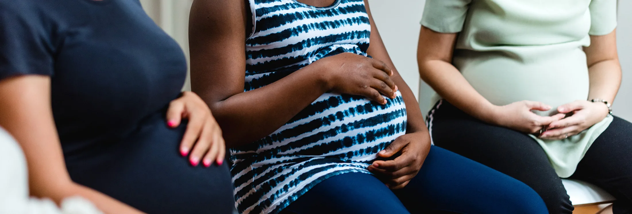 Three pregnant women sit side by side, each resting a hand on their baby bumps. Their faces are not visible, and they are wearing comfortable clothing in different colors and patterns.