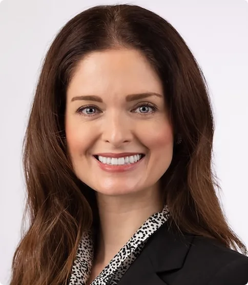 A woman with long brown hair, wearing a black blazer and a patterned blouse, smiles at the camera against a plain light background.