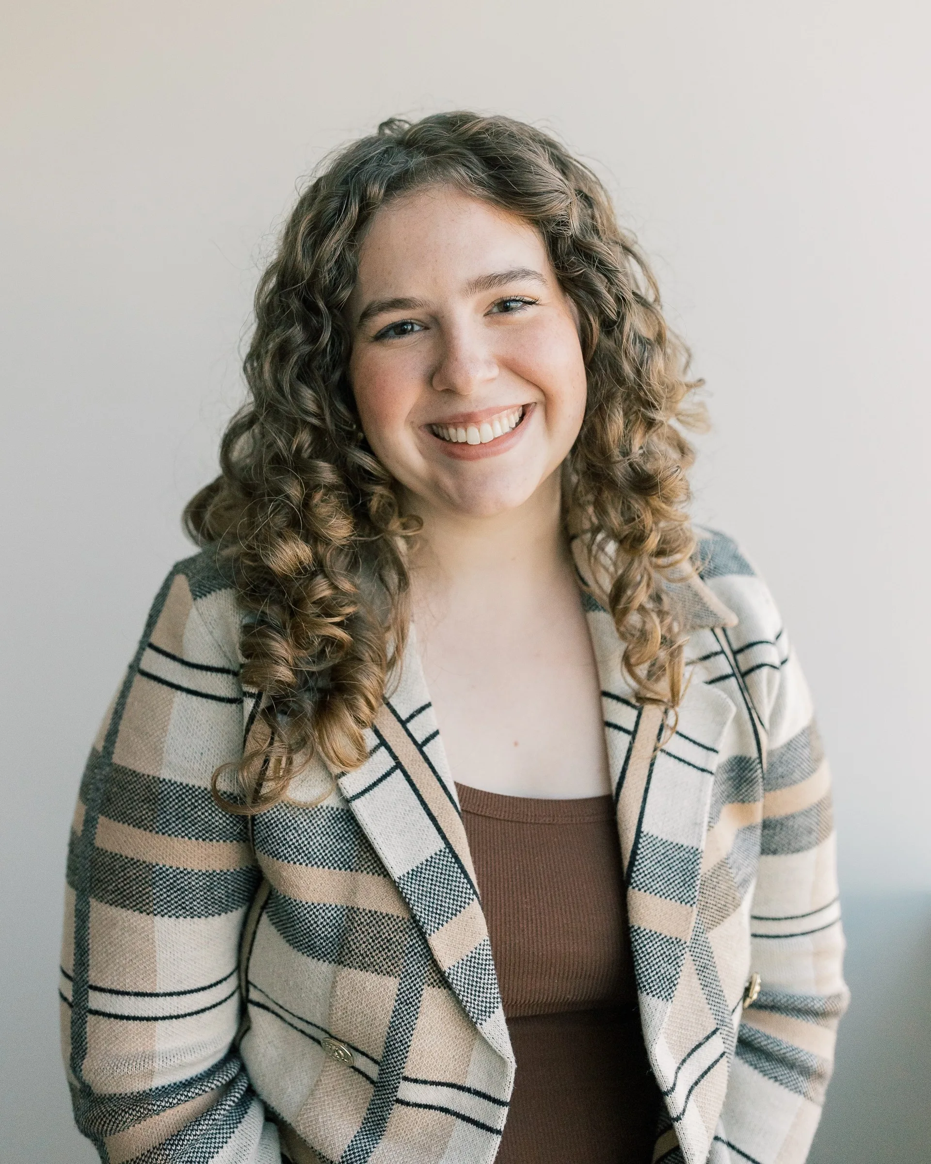 A woman with curly brown hair smiles at the camera, wearing a plaid blazer over a brown top, standing in front of a plain light-colored background.