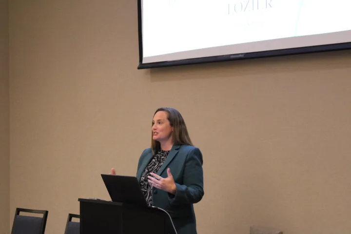 A woman in a green blazer speaks at a podium with a laptop, gesturing with her hands as she addresses the importance of building a pro-life safety net. An empty chair and a projection screen are visible in the background.