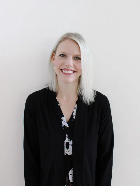 Smiling woman with straight, shoulder-length blonde hair wearing a black jacket over a patterned blouse, standing in front of a plain white background, embodying the spirit of a strong pro-life safety net advocate.