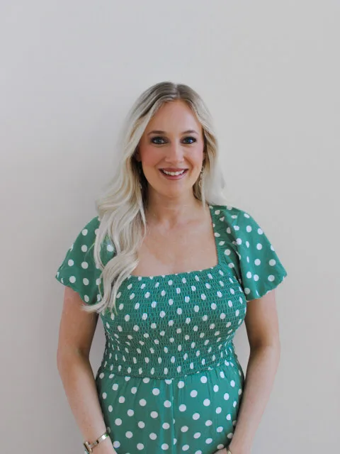 A smiling woman with long blonde hair wearing a green dress with white polka dots stands in front of a plain light-colored wall, embodying the warmth and reassurance of a pro-life safety net.
