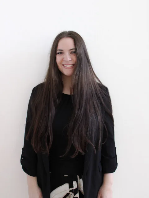 A woman with long brown hair, wearing a black top and jacket, stands smiling against a plain white background.