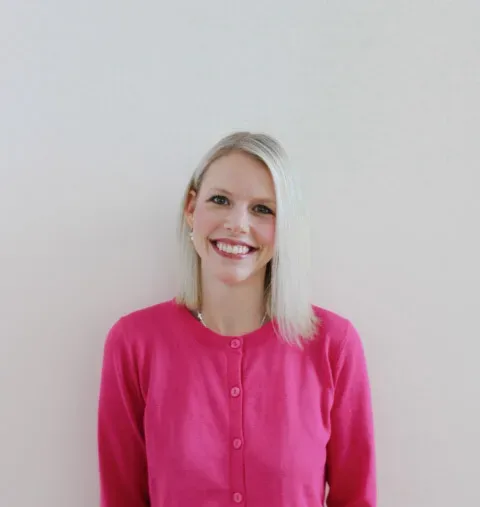 A woman with straight blonde hair, wearing a bright pink cardigan, stands smiling against a plain light-colored background.