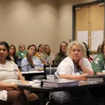 Several women sit at tables in a classroom or conference room, attentively listening to a speaker. Notebooks, drinks, and papers are on the tables as they engage in a discussion about strengthening the pro-life safety net. The background shows a closed door and notes posted on the wall.