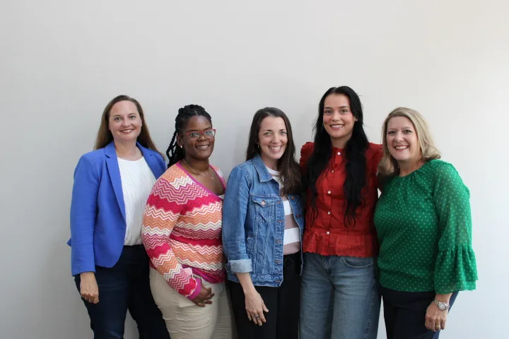 Five women stand together smiling in front of a plain light-colored wall, wearing colorful, casual clothing—including a blue blazer, patterned sweater, denim jacket, red blouse, and green top—reflecting the warmth and camaraderie of a pro-life safety net community.