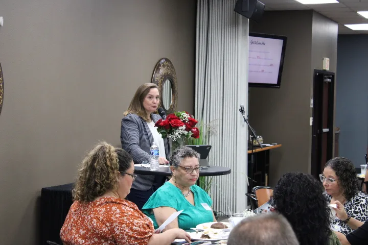 A woman speaks at a podium decorated with red roses to an audience seated at tables.